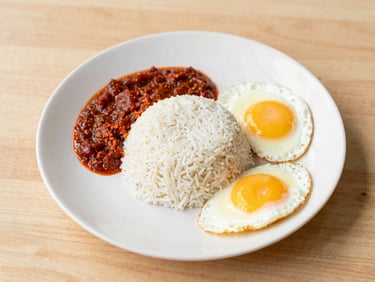 Overhead shot of a full Malaysian breakfast plate with nasi lemak, fried egg, and sambal, on a wooden table, bright natural light, soft yellow accents.