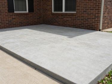 Wide environmental shot of a finished concrete patio adjacent to a brick house in Louisville, smooth surface with clean perimeter edges, natural midday sunlight casting soft shadows, no furniture or people