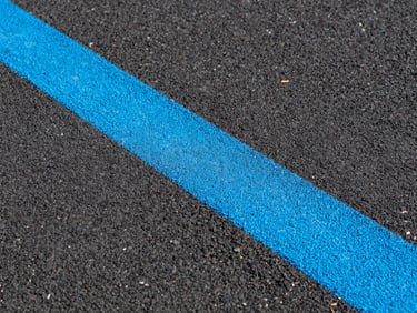 Close-up, low-angle shot of a freshly painted bright blue line on a dark asphalt surface, showing a fine, gritty texture and crisp edges under direct sunlight. Sharp architectural photography, high-contrast vibrant athletic colors.