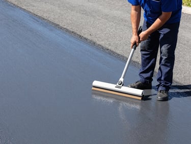 Medium shot of a uniformed operator using a specialized squeegee to apply a dark gray Slurry Seal coating evenly across a wide asphalt surface, with a freshly applied section visible. Bright direct sunlight, crisp details.