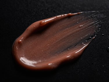 Macro close-up of rich, velvety cosmetics cream texture with wet highlights, low-key dramatic studio lighting, dark obsidian background, 85mm lens