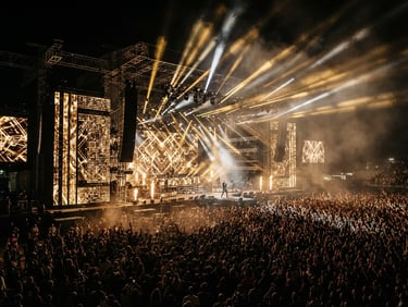 Dramatic wide-angle shot of a massive concert stage at night, glowing golden LED walls, heavy rigging, beams of light cutting through haze over a dense crowd.