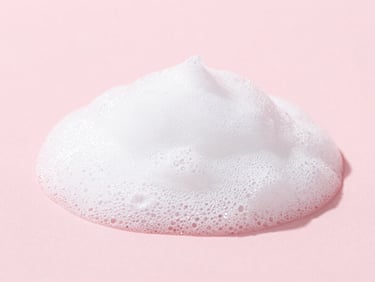 Macro texture close-up of a gentle cleanser foam on a pastel pink background, showing soft light reflection, clean beauty product photography, soft diffused daylight, minimalist composition, 50mm lens.