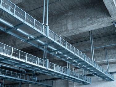 Wide-angle industrial interior showing neatly organized metal cable trays and heavy electrical conduits suspended from a high concrete ceiling, cool blue lighting, crisp technical detail, 35mm