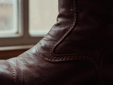 Macro close-up of thick leather edges and clean double-stitching on a dark brown leather boot, moody workshop window light