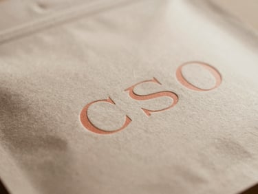 Close-up of premium cosmetic packaging with embossed minimalist typography, tactile paper texture under soft sunlight, muted beige and coral tones, shallow depth of field