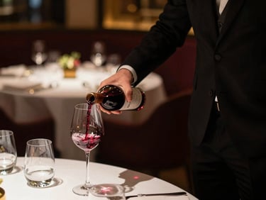 Serveur en costume sombre, main tendue pour verser du vin dans un verre en cristal, éclairage doux et chaleureux, arrière-plan flou d'une salle de restaurant élégante.