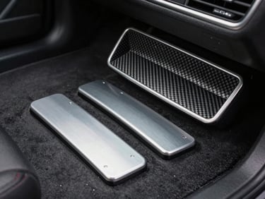 Close-up product shot of interior trim accessories — carbon-look dash insert and aluminum pedal covers — arranged on a dark matte surface under precise studio side-lighting