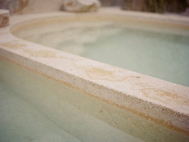 Quiet, unpeopled thermal spa environment, natural textures like travertine, soft golden light, muted beige tones, shallow depth of field, 35mm film style