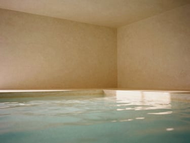 Wide shot of a minimalist, serene subterranean thermal spa interior with natural light filtering in, warm alabaster tones, and tranquil water
