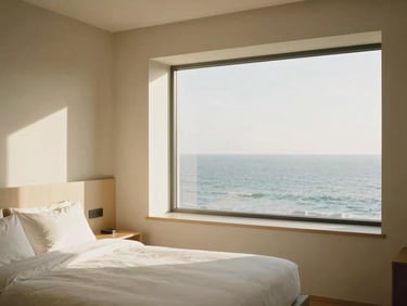 Interior of a minimalist luxury hotel suite, sun-drenched, alabaster and sand tones, large window overlooking the ocean, unpeopled, warm editorial photography, 35mm film style