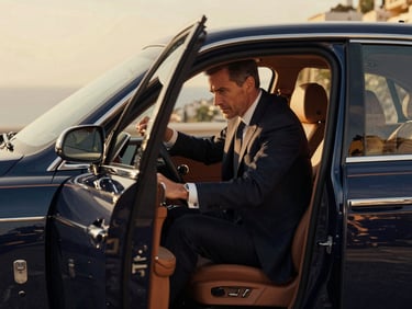 A professional driver in a tailored dark suit opening the door of a pristine navy luxury vehicle, warm Mediterranean sunlight