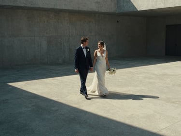 Cinematic wide shot of a bride and groom walking through a minimalist concrete courtyard, harsh side lighting casting long shadows, 35mm film grain, muted tones.
