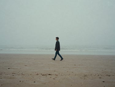 Cinematic 35mm still of a lone figure walking on a foggy beach, muted blue and gray tones, wide landscape framing, minimalist composition.