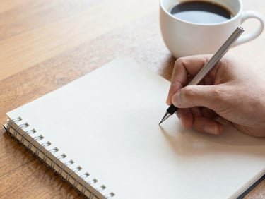 Close-up of a hand writing on a clean notebook next to a cup of coffee on a wooden table, bright natural light, transparent and organized atmosphere