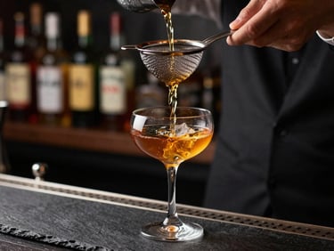 Bartending station close-up — a cocktail being poured through a strainer into a coupe glass, amber liquid catching overhead bar light against a dark slate surface, bottles blurred in background