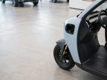 Wide overhead shot of an EV showroom floor — front wheel and charging port of an electric three-wheeler visible on polished concrete, bright clinical white lighting, no people, clean minimal environment