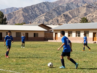 Action shot of high school boys playing soccer on a green field, rustic school buildings and Andes mountains in the background, bright daylight.