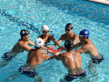 Action shot of lifeguards in training, executing a synchronized rescue drill in a deep pool, bright daylight casting sharp shadows on the water surface.