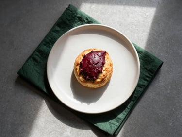 Overhead flat-lay of a minimalist Scandinavian table setting, hand-thrown ceramic plate with a single artisanal pastry, soft morning window light, high contrast shadows, deep forest green napkin.