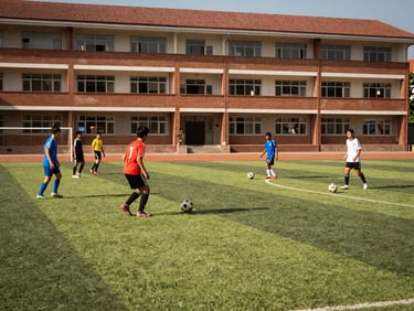 Students playing soccer on the school field, bright natural daylight, wide-angle shot, school building in the background, rich deep tones.