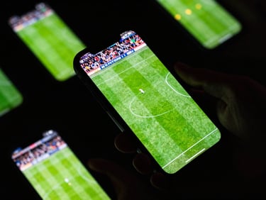 Close-up shot of a smartphone screen displaying a live football match with vibrant green grass, held by hands in stadium stands, high-contrast neon lighting.