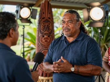 A professional broadcast interviewer speaking with a Samoan high chief under a traditional fale, professional lighting setup.