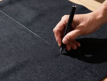 Close-up of a tailor's hands chalking a pattern on heavy charcoal wool, crisp studio lighting highlighting fabric texture.
