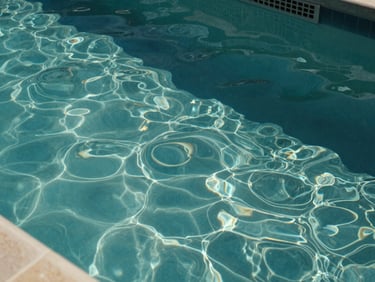 Overhead close-up of clear residential pool water, light refracting on the bottom tiles, pool skimmer basket visible at the edge, mid-morning natural light, no people