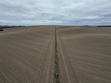 Wide-angle drone shot of rolling vacant fields divided by a straight wire fence under a cool grey overcast sky, technical framing.