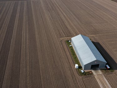 High-detail aerial view of structured agricultural crop fields, a single metal barn on the right, desaturated earth tones.