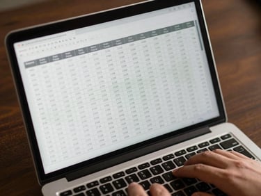 Close overhead view of a structured financial model on a laptop screen, columns of figures clearly organized, a hand resting on a keyboard at the lower right, warm tungsten office light reflecting off the screen surface, dark desk background