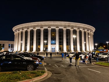 Wide environmental shot of The Forum's iconic circular exterior at night, event lighting spilling onto the parking perimeter, crowd moving toward curb pickup zones, real ambient streetlight, dynamic angle from ground level