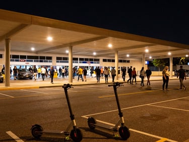 Wide environmental shot of Kia Forum parking perimeter at night, crowd streaming from the arena exit toward the street, overhead parking lot lights casting real amber glow, electric scooters staged at the curb in the foreground, dynamic angle from street level