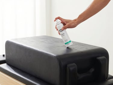 Hands applying premium Inova Protect spray onto a sleek black leather Pilates carriage, bright daylight, 50mm