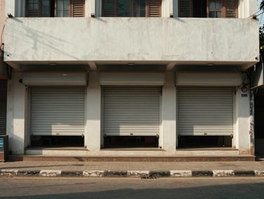 Street-level exterior of a single-storey commercial building in Chennai, midday overhead light, shopfront shutters open, plain rendered facade, road and footpath in foreground