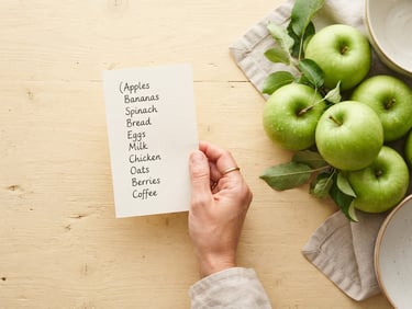 Bright high-key photography of a hand holding a clean handwritten grocery list next to fresh green apples on a warm cream table.