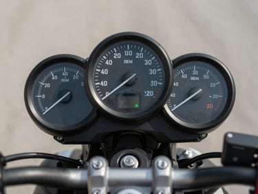 Close-up overhead shot of a motorcycle fuel gauge and speedometer cluster on a neutral studio surface, instrument dial markings visible under soft daylight, no rider or background clutter