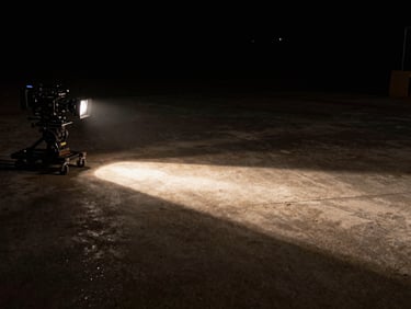 Empty film set at night, practical tungsten fresnel casting a hard diagonal shaft across a dark concrete floor, camera on a dolly in the left third, deep background falling off to black, cinematic medium wide shot