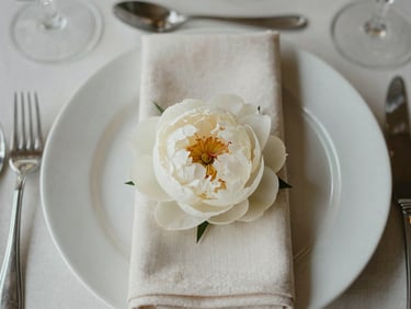 Close-up overhead of a finished wedding table arrangement — folded ivory napkins, crystal glassware, a single white peony at centre, soft diffused overcast light, linen texture visible