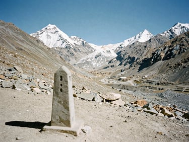Wide-angle rugged alpine landscape of a high Himalayan pass under harsh daylight, desaturated tones, showing a simple stone trail marker in the foreground, 35mm film.