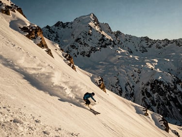 Wide environmental shot of a skier carving through deep powder on a steep alpine face, dramatic golden hour light on peaks.