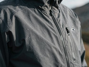 Extreme close-up of a technical hardshell jacket fabric with water droplets, sharp focus, desaturated alpine tones.