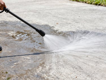 Pressure washer spray cleaning a pale concrete driveway on a bright Florida noon, water dispersing across the surface with clean half visible, dark weathered half in contrast