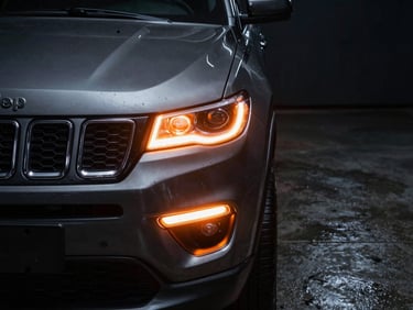 Dramatic close-up of a dark grey Jeep Compass SUV headlight and grille, intense orange light glow from the side, dark wet concrete floor, cinematic, 35mm