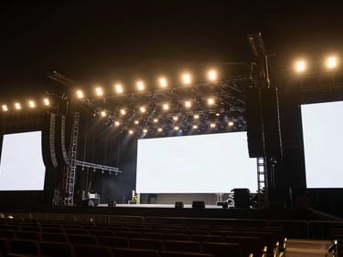 Wide shot of a large, professionally lit concert stage with complex trussing and LED screens, empty before an event, cinematic event photography, dramatic ambient lighting, rich dark tones with gold accents, shallow depth of field, 35mm