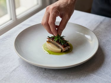 Close overhead of a carefully plated dish on a matte ceramic plate, natural window light from the left, a chef's hand adjusting a herb garnish, Nordic-inspired clean table linen underneath, shallow depth of field