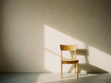 A minimalist photography studio interior with soft natural window light casting gentle shadows on a neutral warm-sand wall, a single oak chair, 35mm film aesthetic.