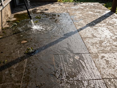 Wide environmental shot, afternoon sun, a stone patio — left half dark with algae and winter grime, right half gleaming clean after high-pressure cleaning. Water streams visible on the clean side. No furniture, no people, purely documentary.