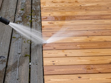 Close medium shot, bright natural daylight, a wooden decking surface — left portion visibly grey and mossy, right portion freshly cleaned to warm natural wood tone. High-pressure water jet mid-action at the transition line. No people, no props.